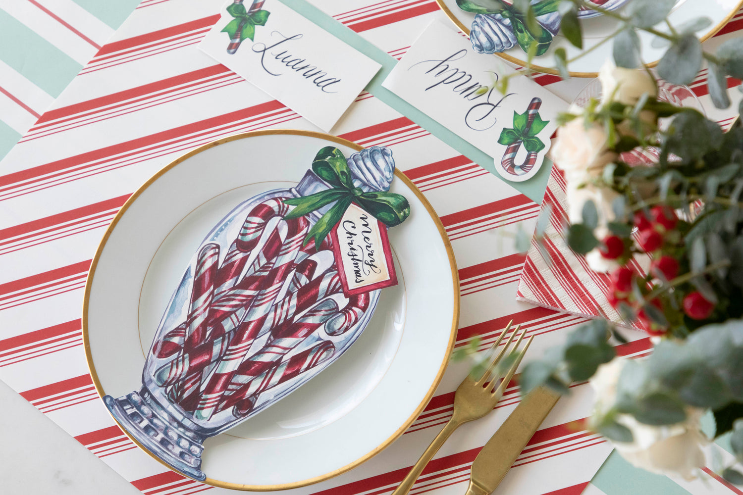 An elegant table setting with the Candy Cane Jar Table accent with &quot;Merry Christmas&quot; written on the tag, on top of a gold rimmed plate that sits on the Peppermint Stripe Placemat.