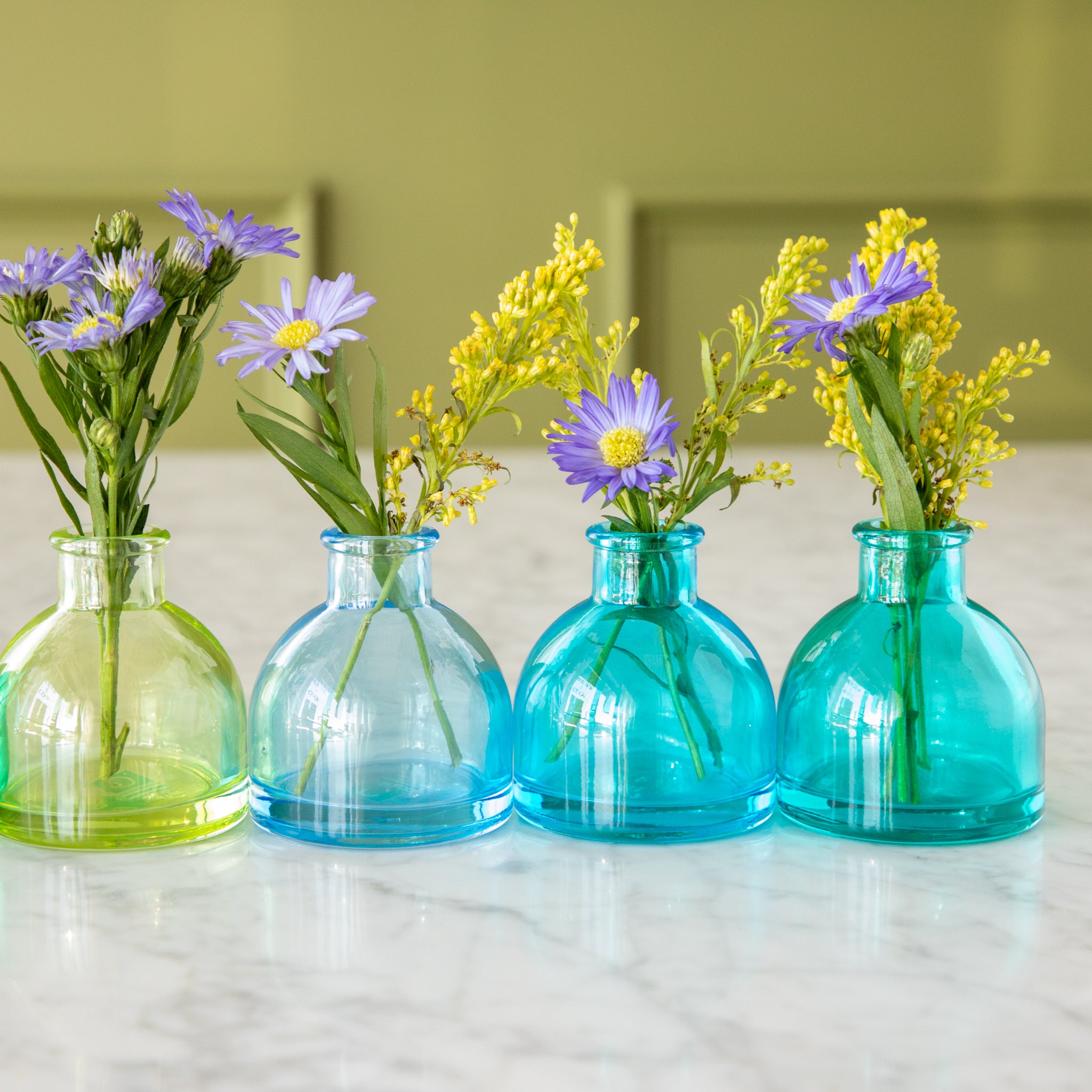 Four round Jewel Tone Glass Bud Vases in green to blue colors with flowers in them.