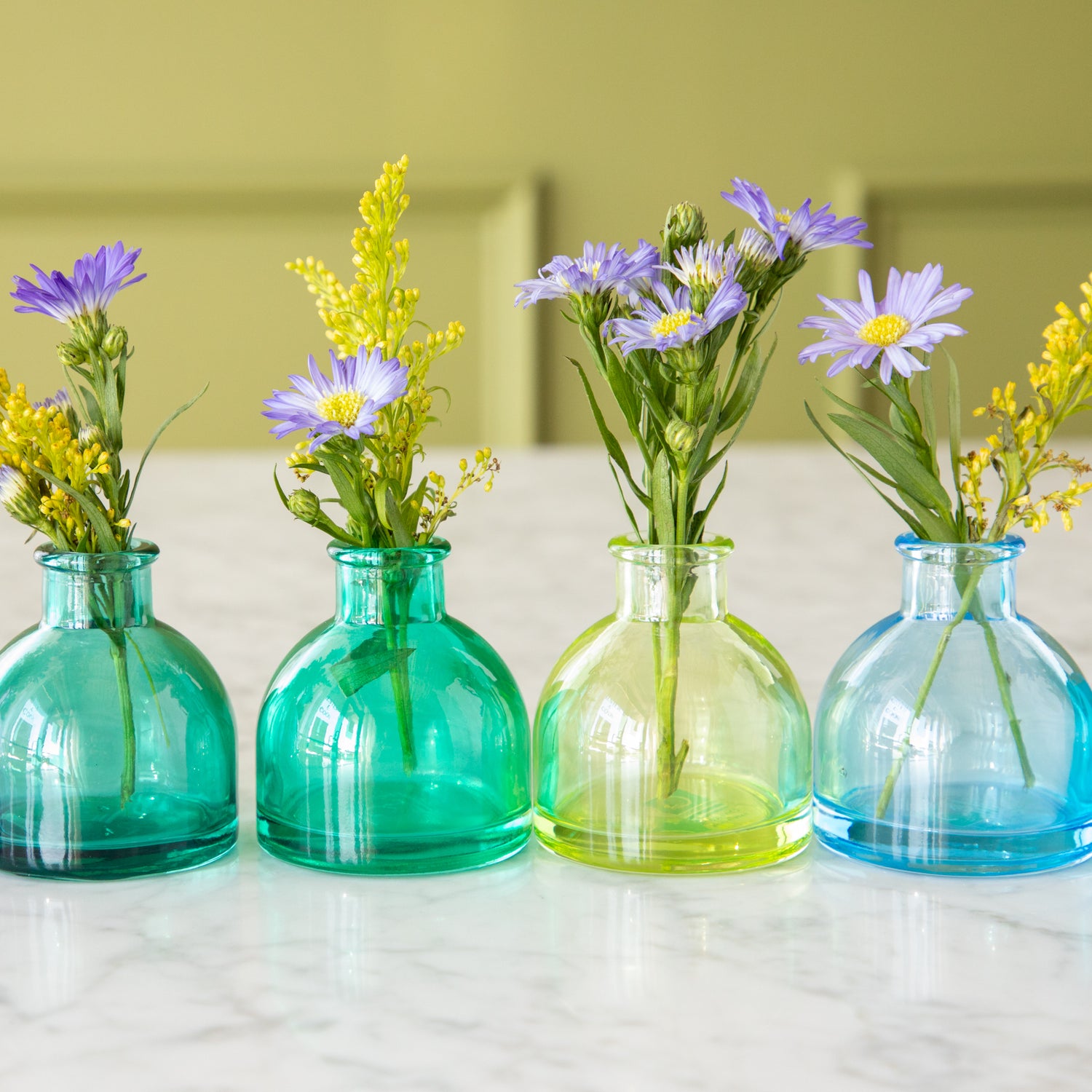 Four round Jewel Tone Glass Bud Vases in green to blue colors with flowers in them.