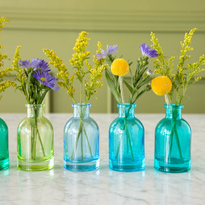 Four Tall Jewel Tone Glass Bud Vases in green to blue colors with flowers in them.