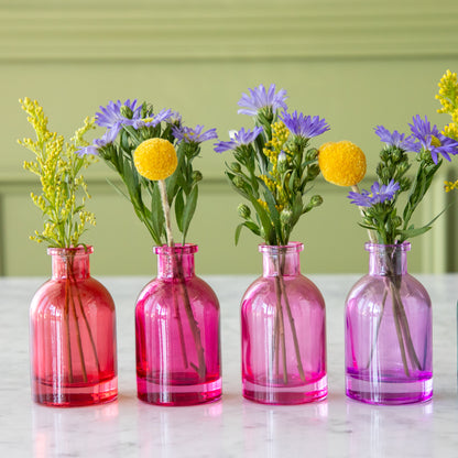 Four Tall Jewel Tone Glass Bud Vases in red to purple colors with flowers in them.