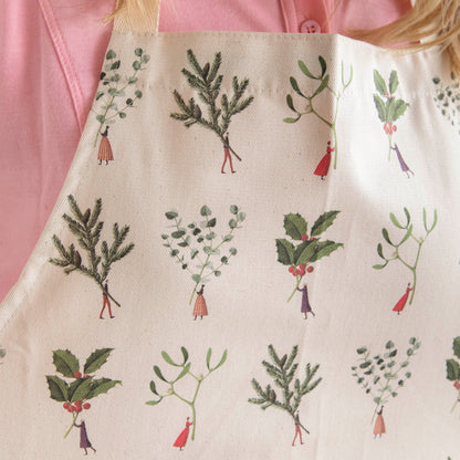 A close up of the Laura Stoddart Christmas apron patterned with illustrated tiny people holding gigantic winter botanicals by the stems.