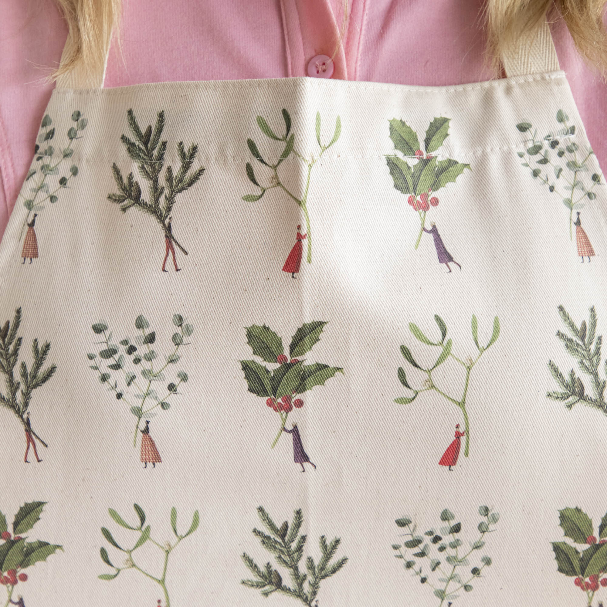 A close up of the Laura Stoddart Christmas apron patterned with illustrated tiny people holding gigantic winter botanicals by the stems.