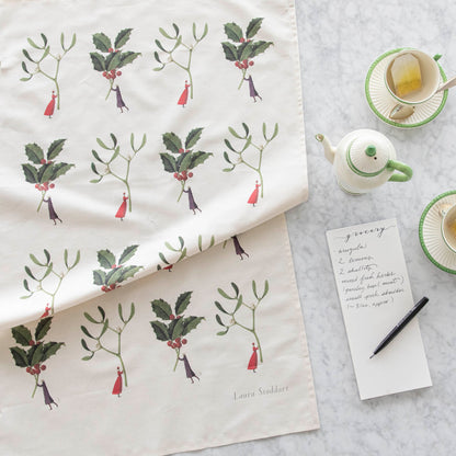 Laura Stoddart Holly &amp; Mistletoe Tea Towel next to a notepad with a grocery list written on it,  tea cups and a creamer pot.