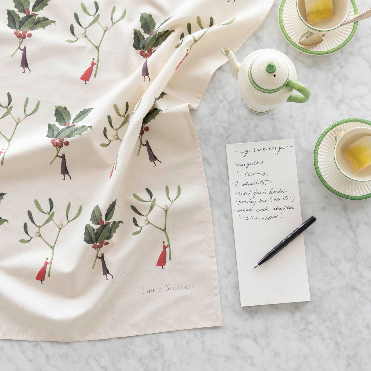 Laura Stoddart Holly &amp; Mistletoe Tea Towel next to a notepad with a grocery list written on it, tea cups and a creamer pot.