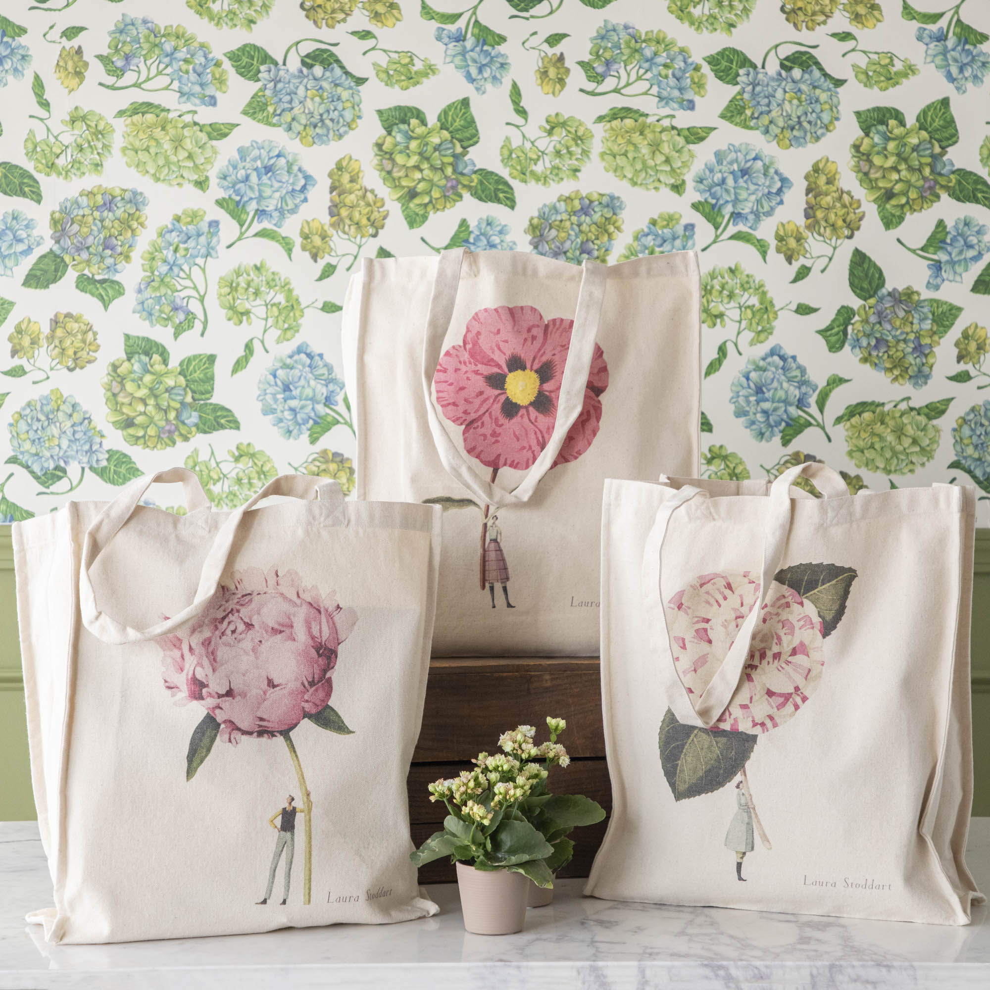 Three of the Heavyweight Canvas Tote Bags featuring pink floral artwork by Laura Stoddart arranged alongside potted plants against a hydrangea wallpaper background.