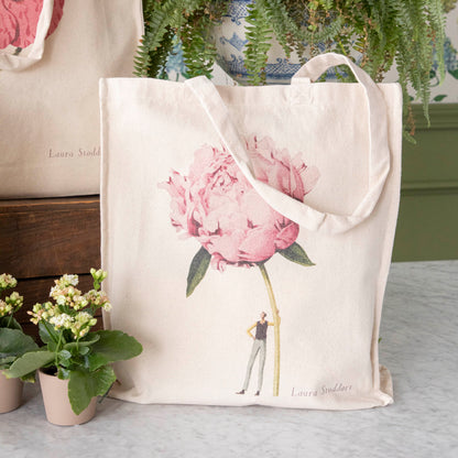 Close-up of the Pink Peony Heavyweight Canvas Tote Bag standing upright in a display, showing the beautiful illustration of a man holding a gigantic pink flower.