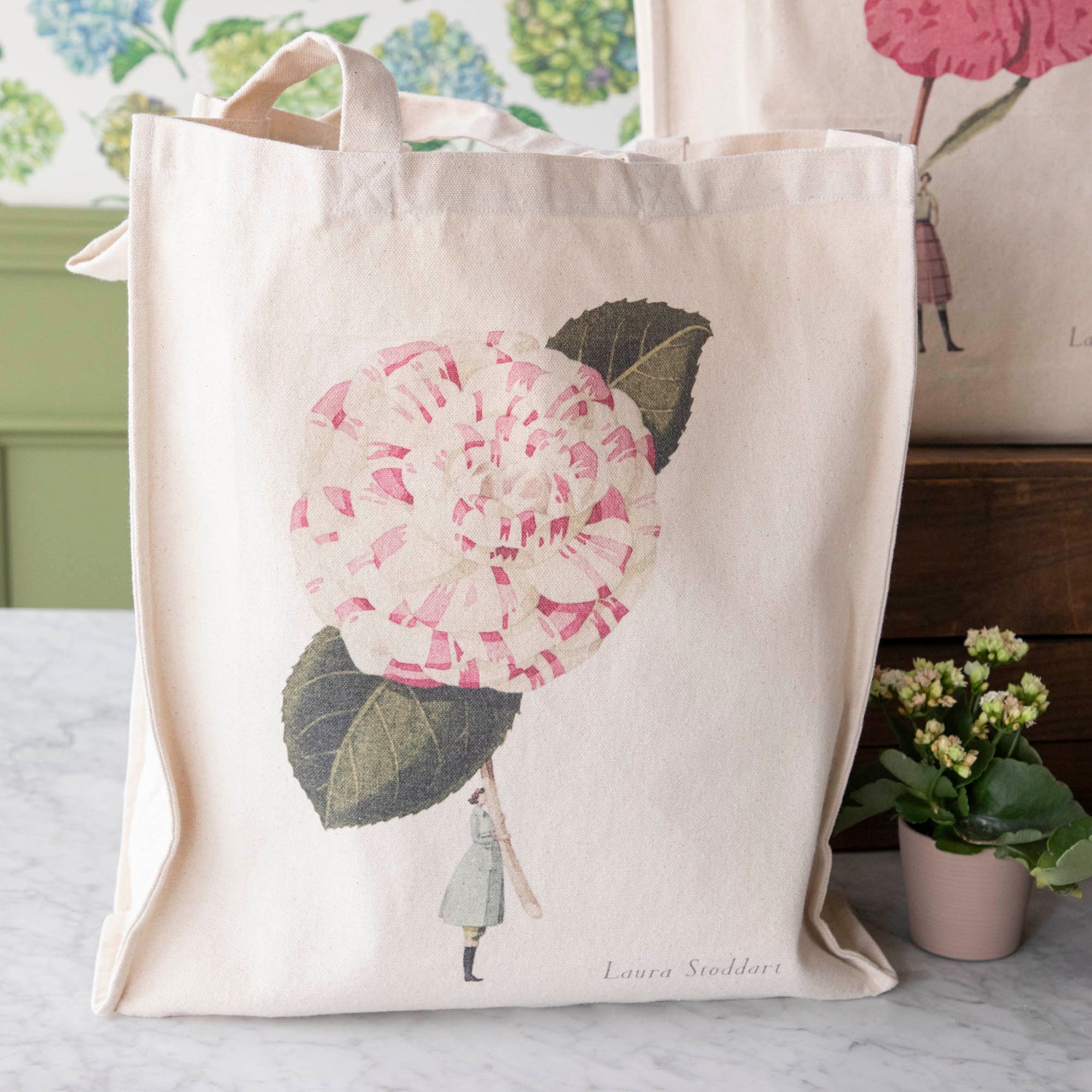 Close-up of the Camelia Heavyweight Canvas Tote Bag standing upright in a display, showing the beautiful illustration of a woman holding a gigantic white and pink flower by the stem.