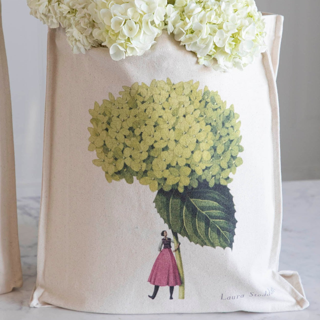 Close-up of the Annabelle Heavyweight Canvas Tote Bag standing upright in a display, showing the beautiful illustration of a woman holding a gigantic green hydrangea bloom by the stem.