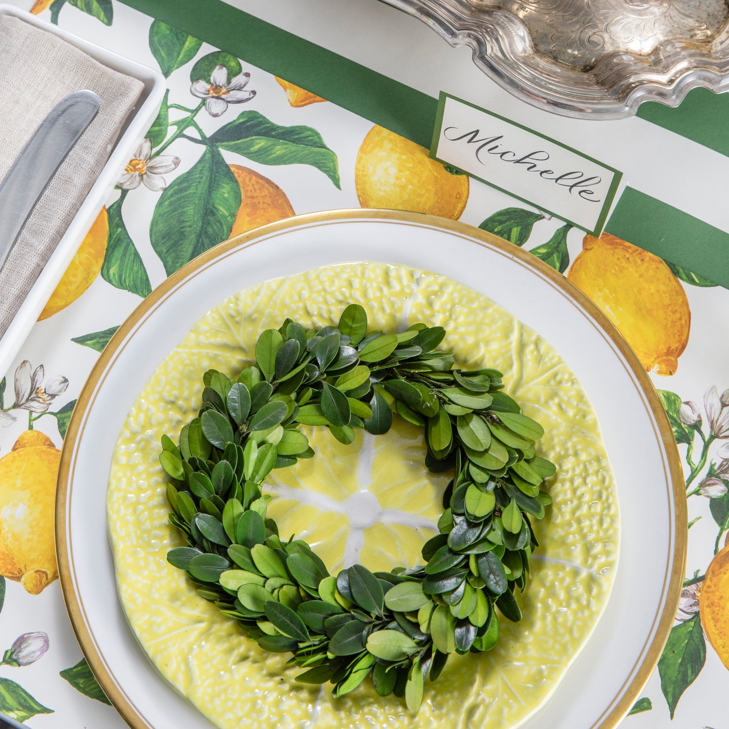 Mini boxwood wreath laying on a lemon shaped plate with a lemon printed placemat below. 