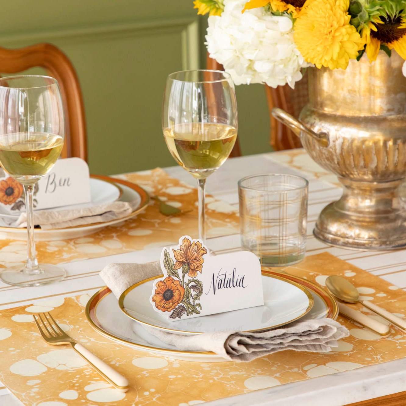 An elegant fall themed table setting with the Gold Stone Marbled Placemat, orange and gold rimmed dinner plates with the Autumn Arrangement place card with the name &quot;Natalia&quot; written on it, on top. 