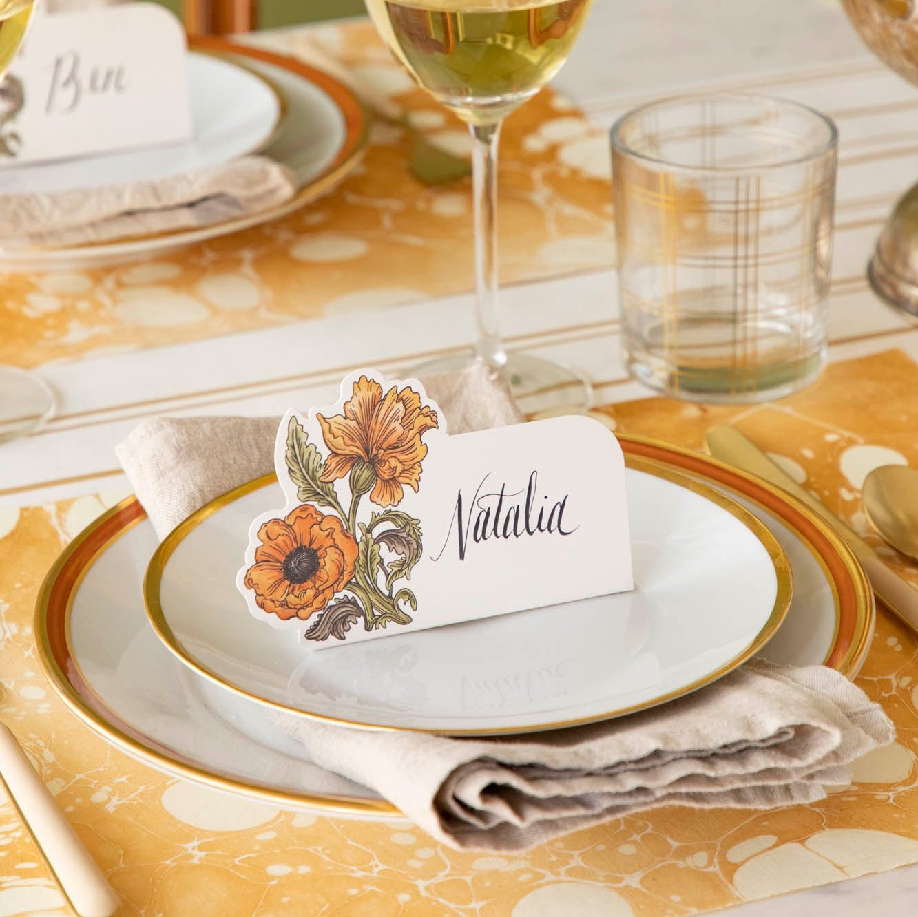 The Autumn Arrangement Place Card with the name &quot;Natalia&quot; written on it, on an elegant gold themed table setting.