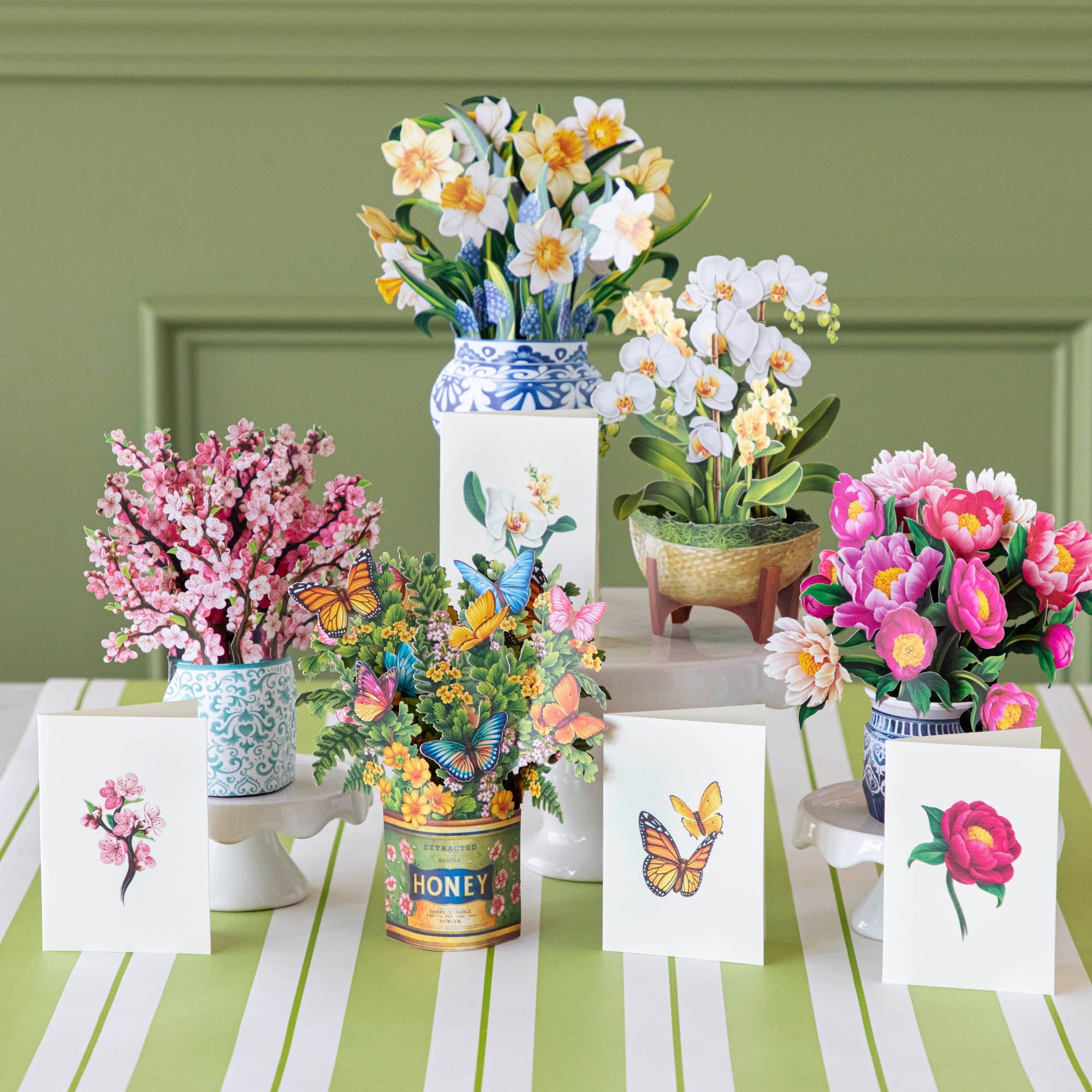 A potted arrangement of artificial orchids with a wooden stand against a white background, accompanied by a Fresh Cut Paper Mini Pop Up Flower Bouquet.