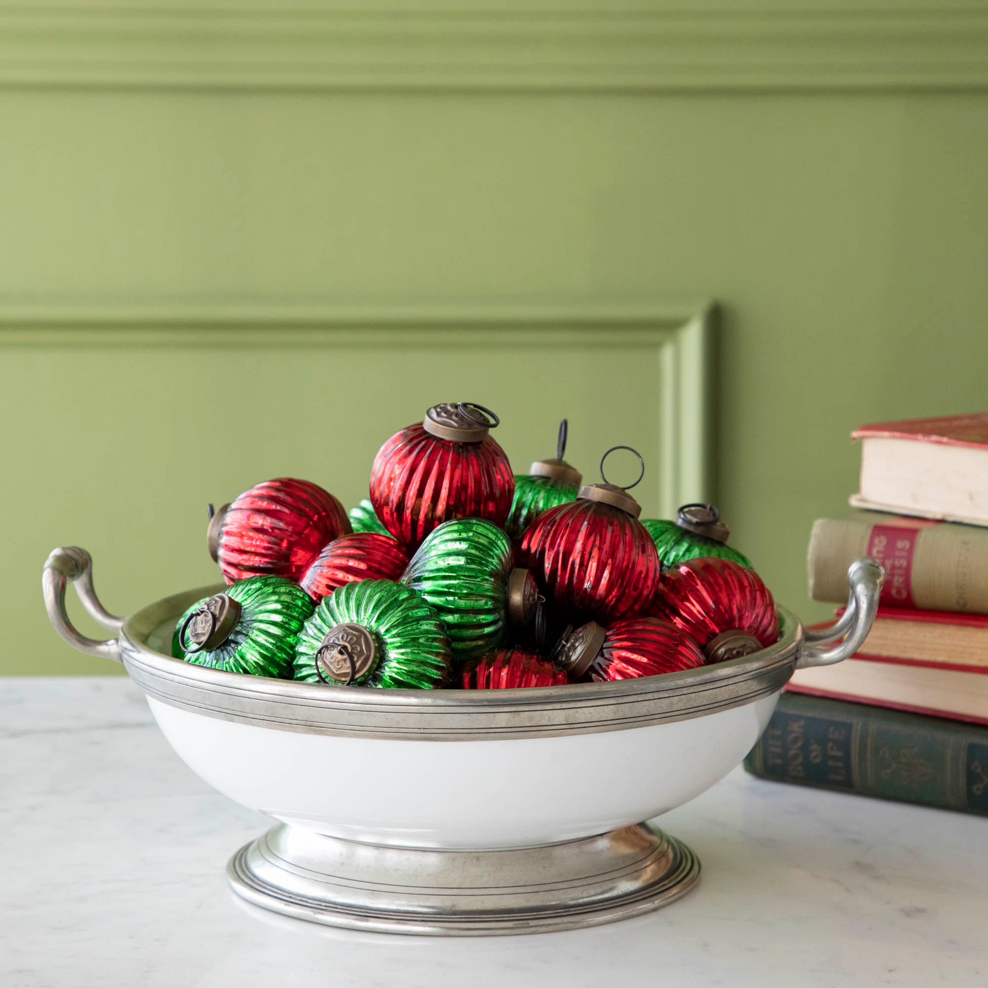 Mini red and green ribbed ornaments decoratively shown in a bowl, in front of a stack of books.