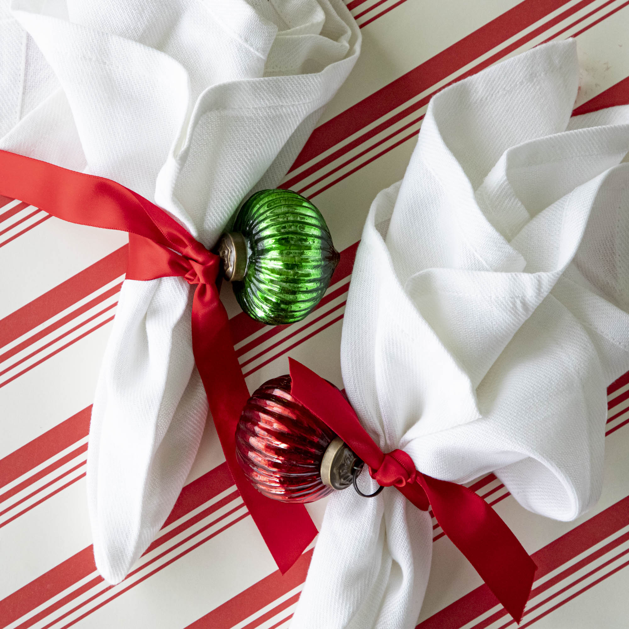 Mini red and green ribbed ornaments tied to a ribbon around a napkin, on a peppermint stripe runner.