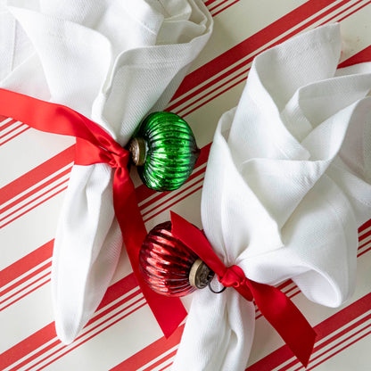 Mini red and green ribbed ornaments tied to a ribbon around a napkin, on a peppermint stripe runner.