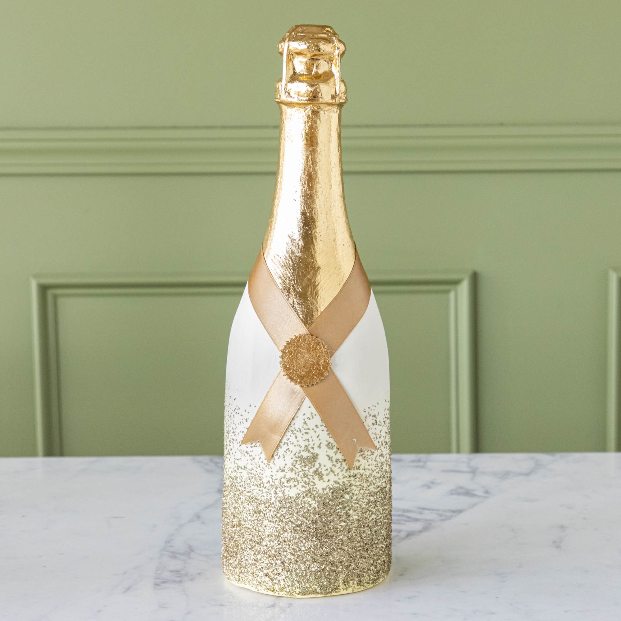 A gold, papermache Oversized Champagne bottle with a gold leafed top, a gold ribbon wrapped around the bottle held together by a gold foil seal, and gold glitter going up the bottle.