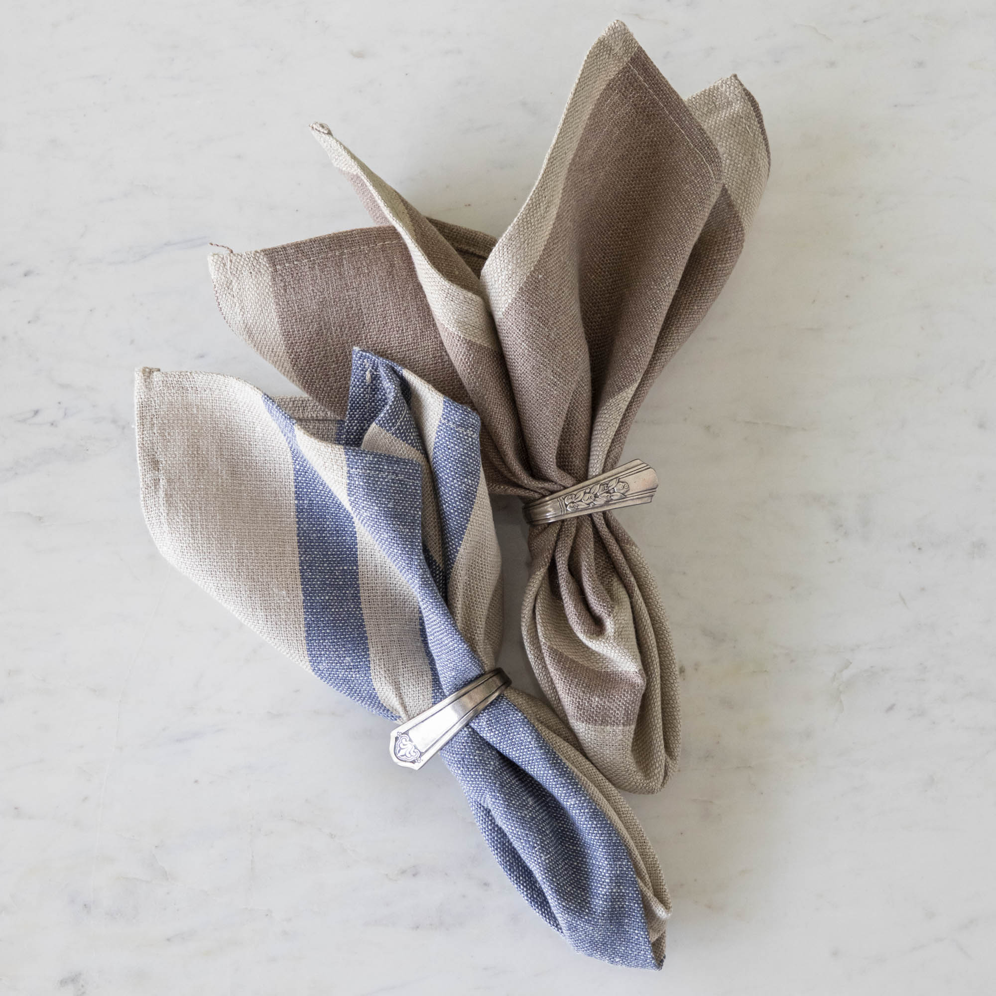 A Vintage Blue Striped Woven Linen Napkin and Dusty Rose Striped Woven Linen Napkin in silver napkin ring holders on a marble table.