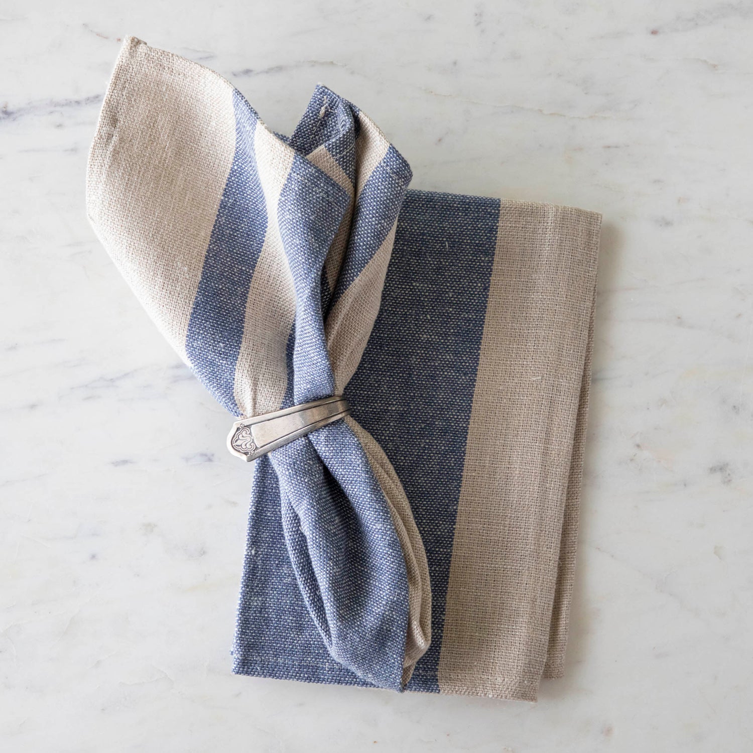 Vintage Blue Striped Woven Linen Napkin in a silver ring napkin holder on top of the same napkin on a marble table.