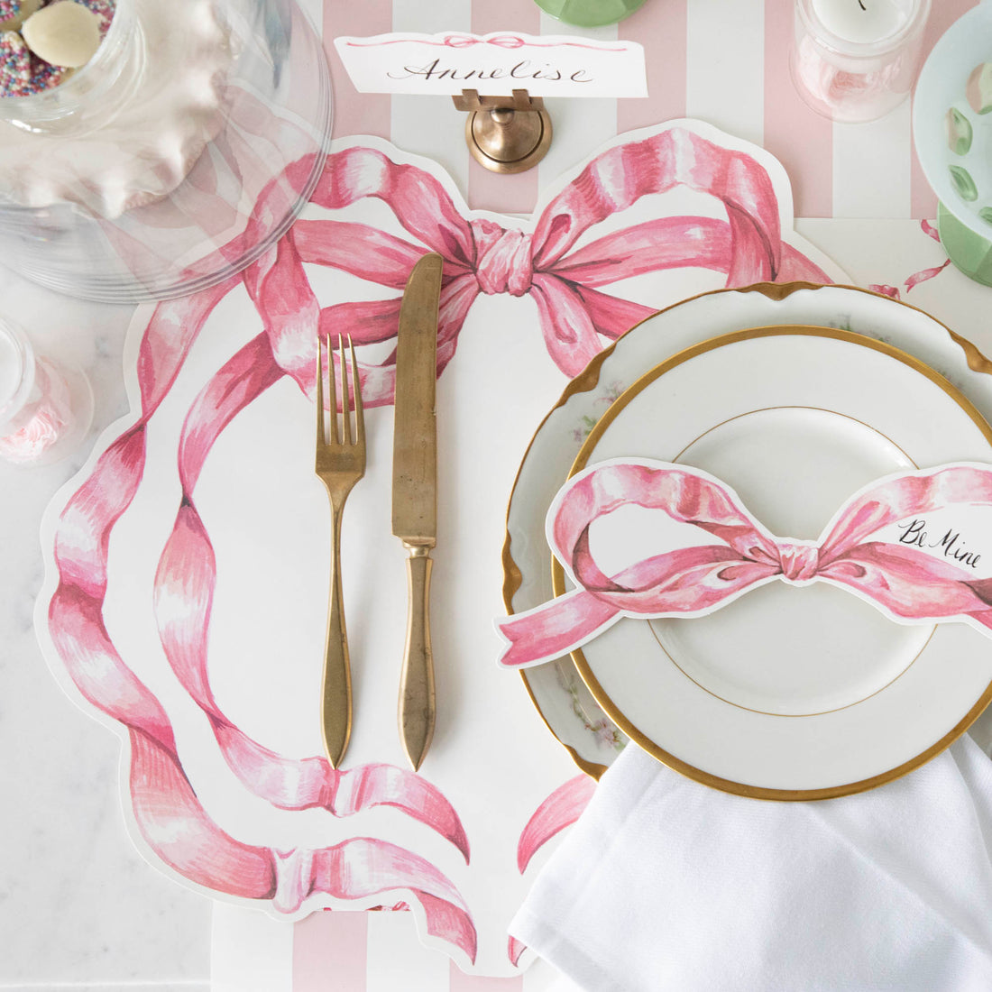 A Pink Bow Table Accent with "Be Mine" written in the blank space in the right bow loop resting on the plate of a pink place setting.