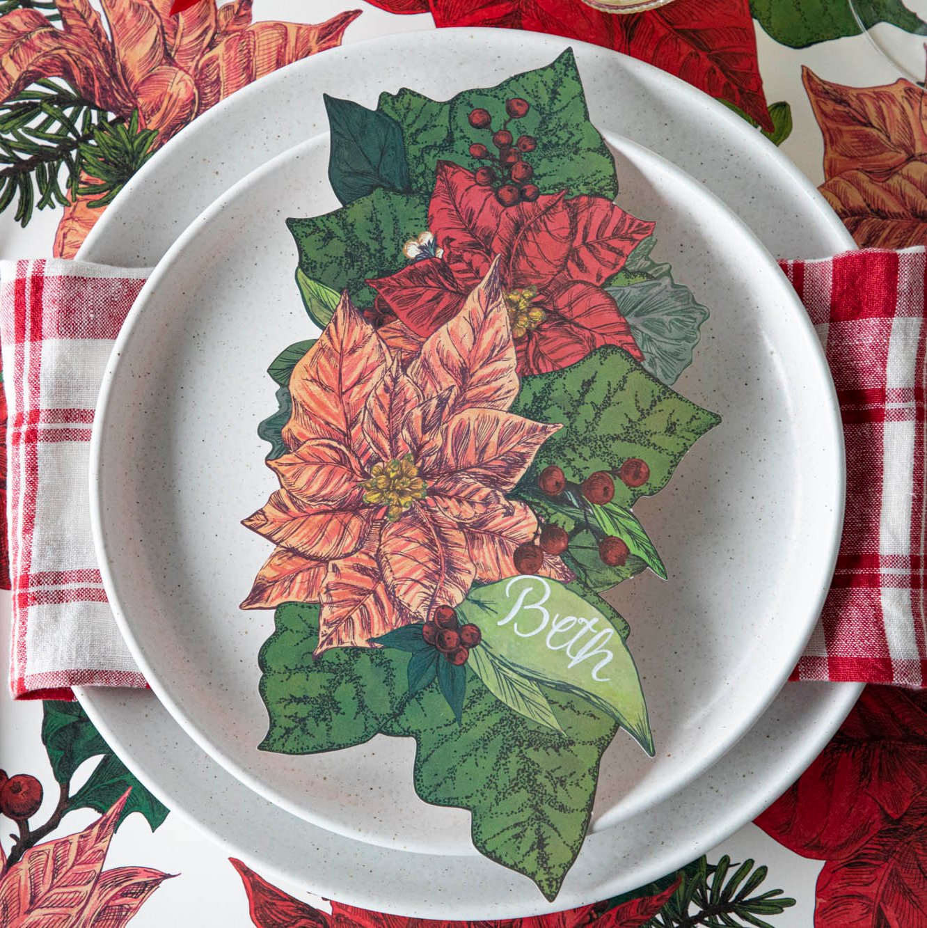 The Poinsettia Table Accent with &quot;Beth&quot; written on the leaf resting on top of Salt Dinner Plates and a red checkered napkin.