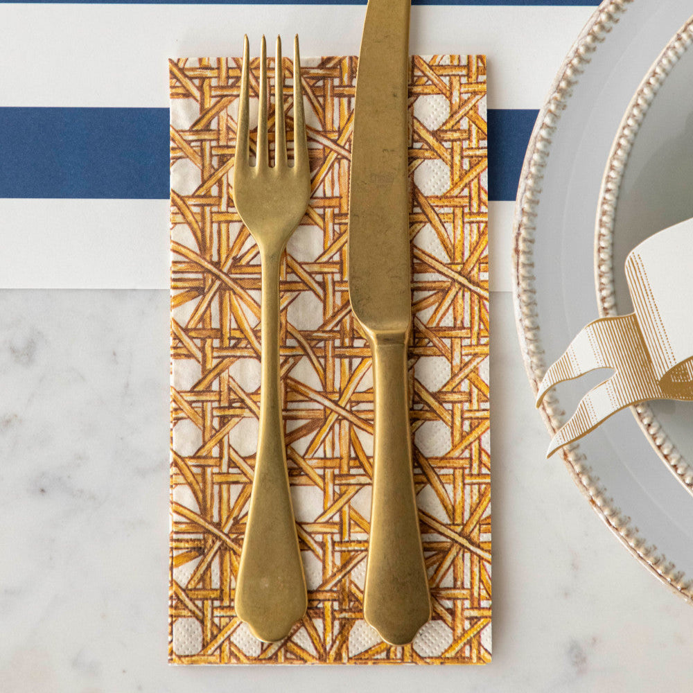 Close-up of a Rattan Weave Guest Napkin under a gold cutlery set next to a plate in an elegant place setting.