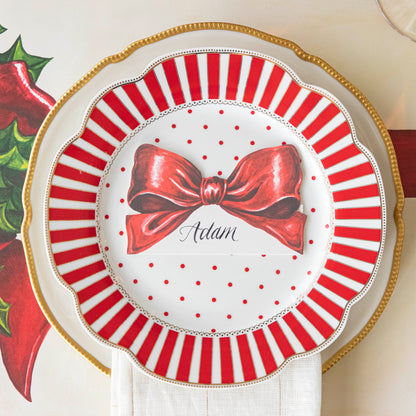 The Bow Place Card with &quot;Adam&quot; written on it in cursive, on a red and white striped plate with red polka dots in the center, on a gold rimmed plate with a white napkin in between.