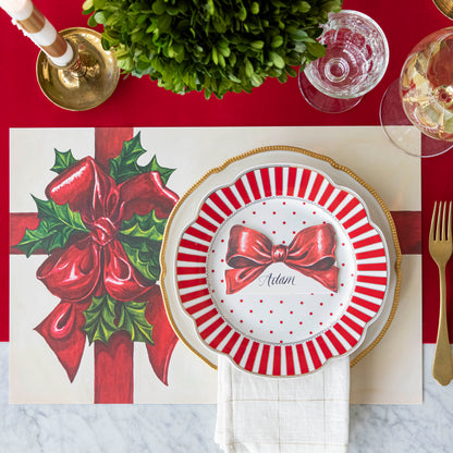 An elegant, Christms themed table setting with the Bow Place Card with &quot;Adam&quot; written on it in cursive, resting on two dinner plates, on top of the Christmas Present Placemat and Red Flocked Runner.