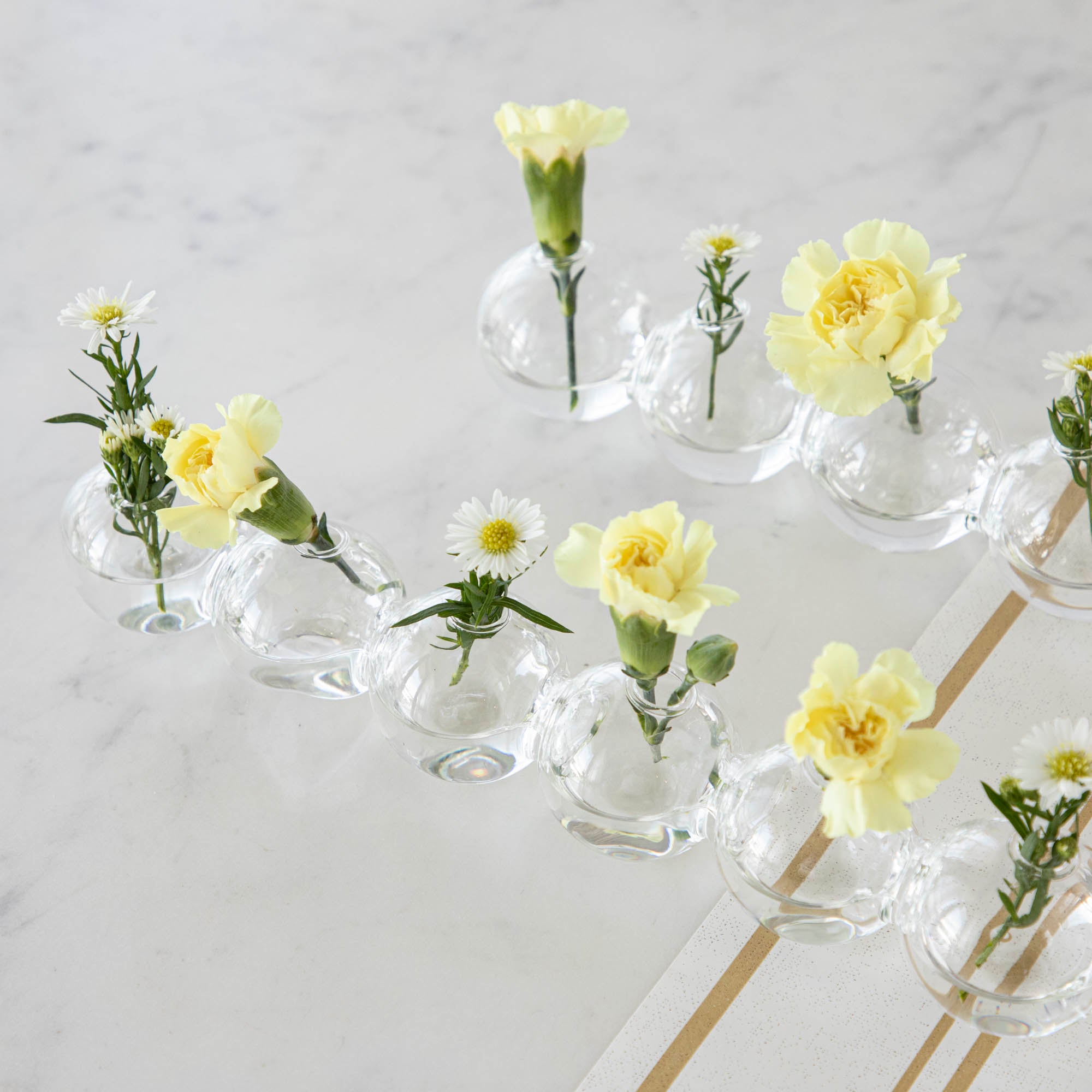 A row of small Chive Caterpillar Vases each containing a single yellow carnation, arranged on a striped surface against a marble background.