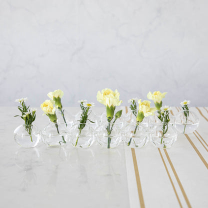 A row of small Chive Caterpillar Vases each containing a single yellow carnation, arranged on a striped surface against a marble background.