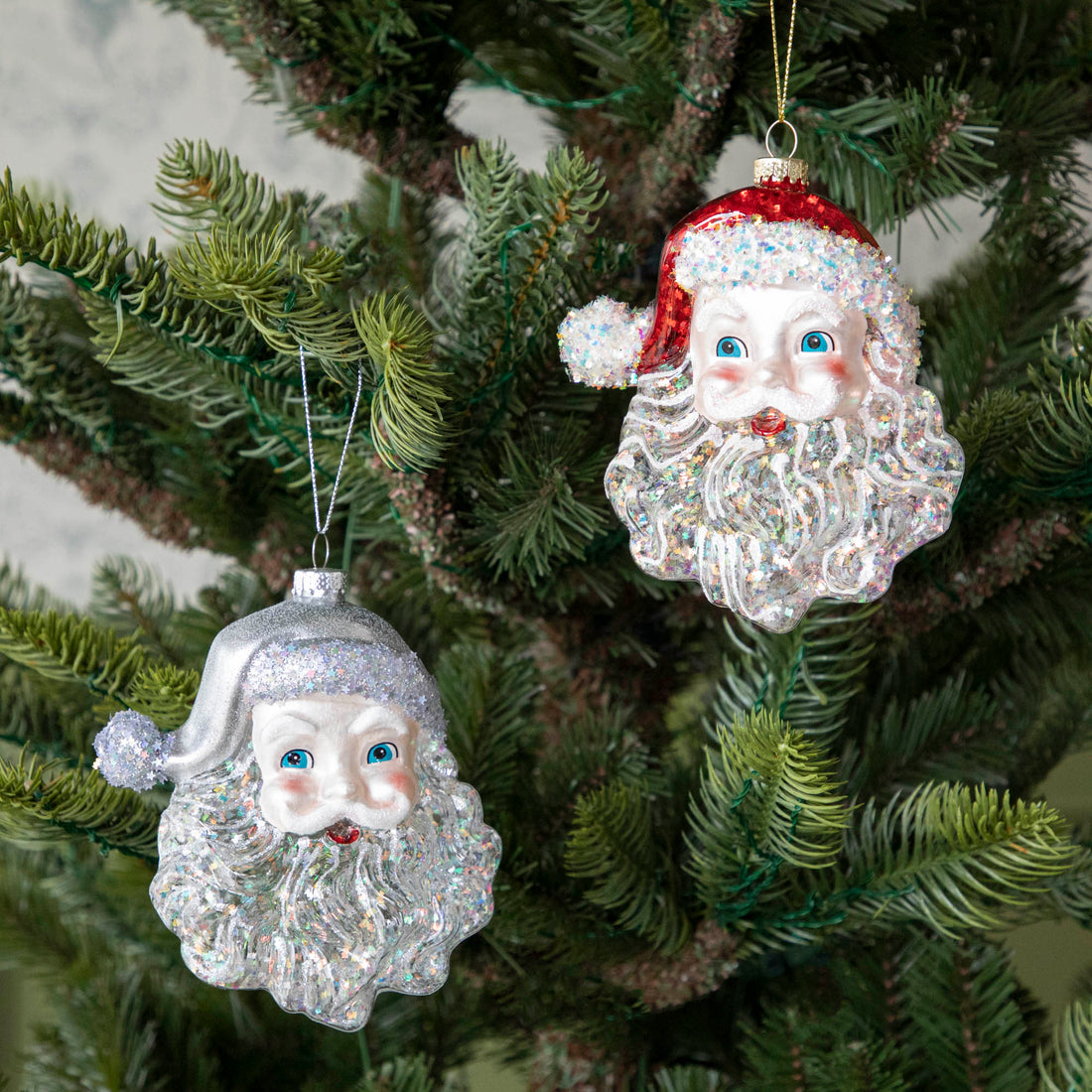 Two Glass Glitter Santa Head Ornaments, one with a silver hat, one with a red hat, hanging on a tree.