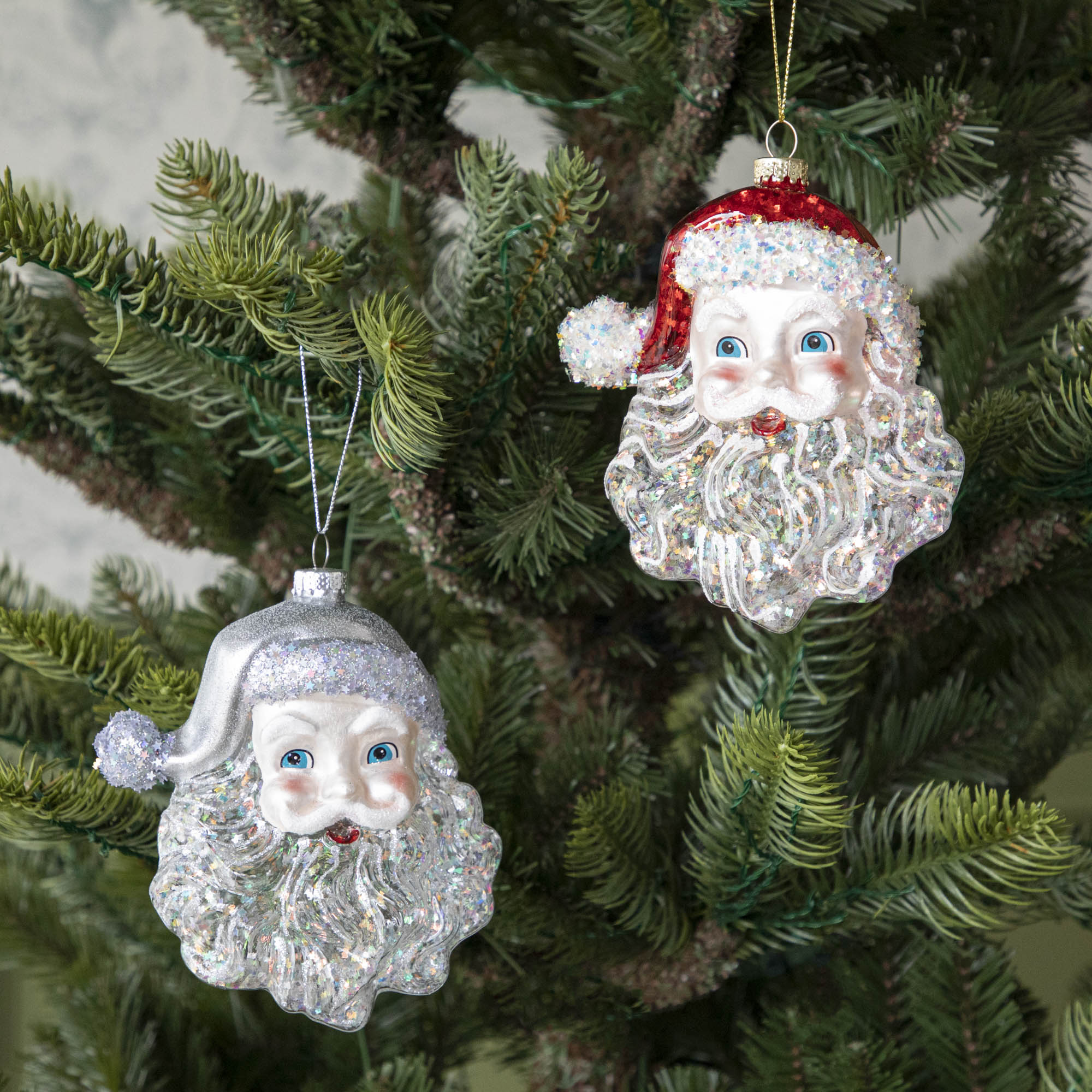 Two Glass Glitter Santa Head Ornaments, one with a silver hat, one with a red hat, hanging on a tree.