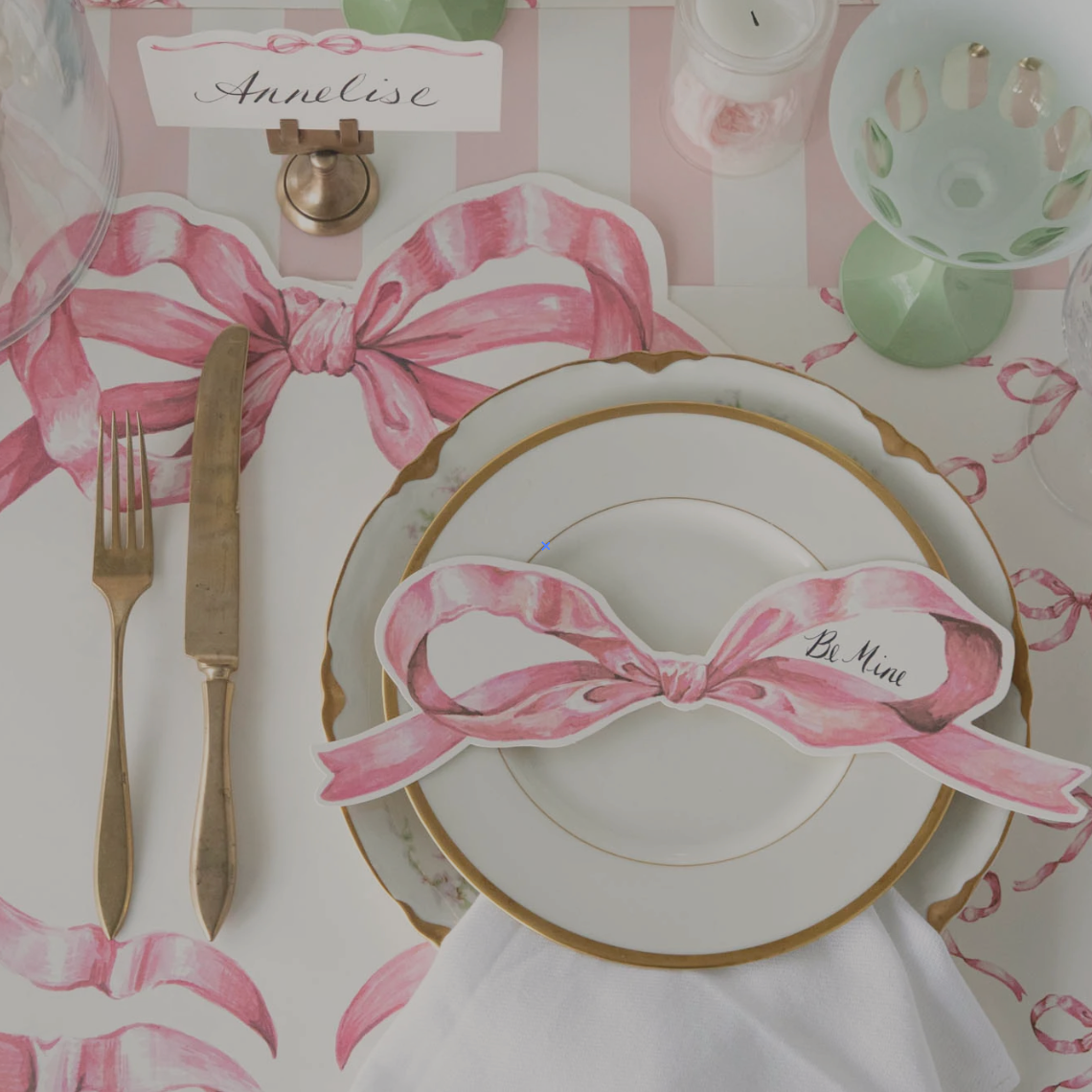 Pink bow placemat on a pink tablesetting with splashes of green and gold silverware. 