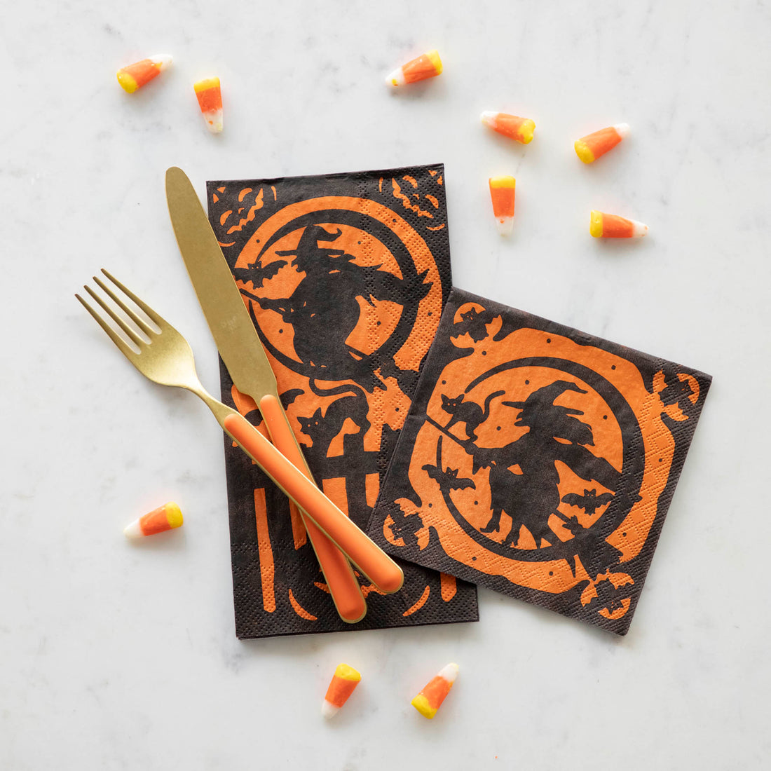 Orange and black wicked witch themed napkins with orange and gold silverware on top, surrounded by candy corn.