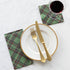 A square cocktail napkin featuring a diagonal plaid pattern of black, red and white over medium green.