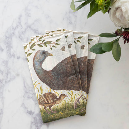 A stack of four Heritage Hen Guest Napkins fanned out on a white table next to a vase of flowers.