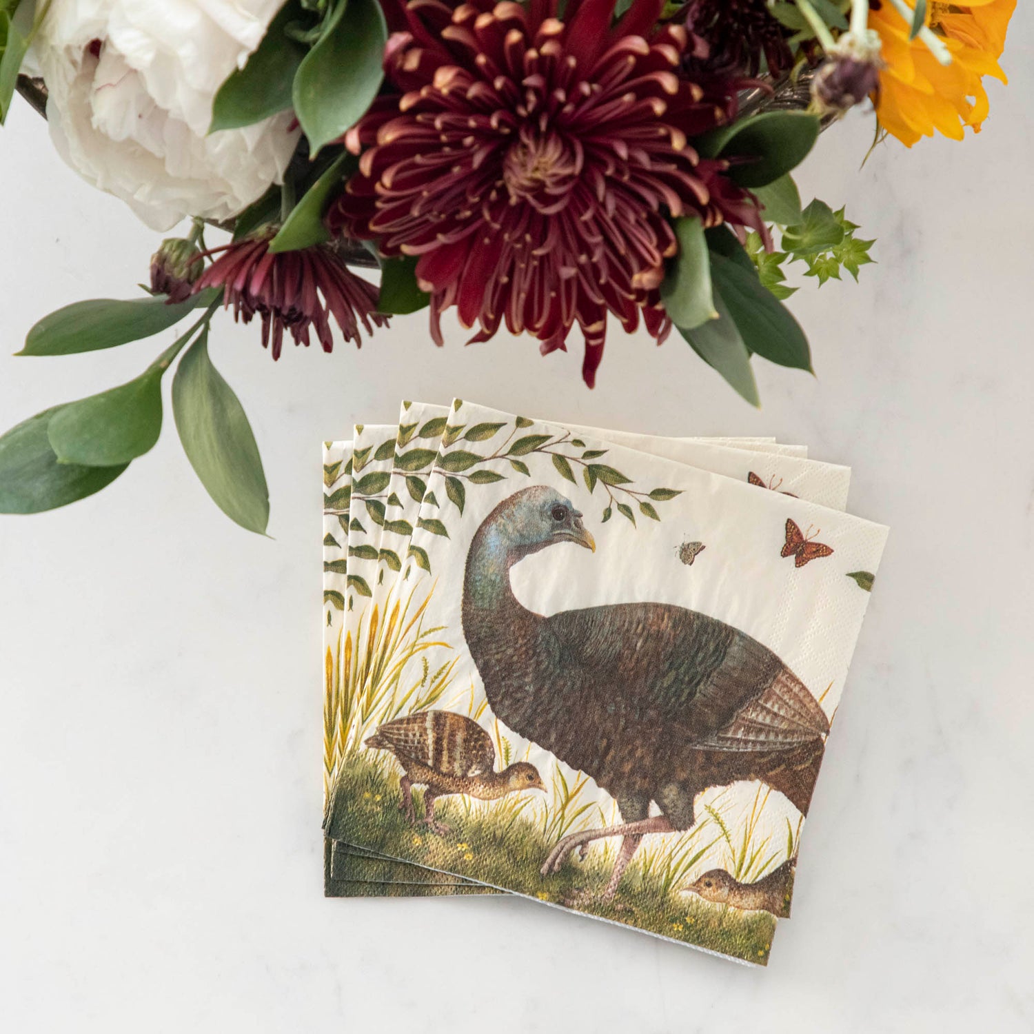 A stack of four Heritage Hen Cocktail Napkins fanned out on a white table next to a vase of flowers.