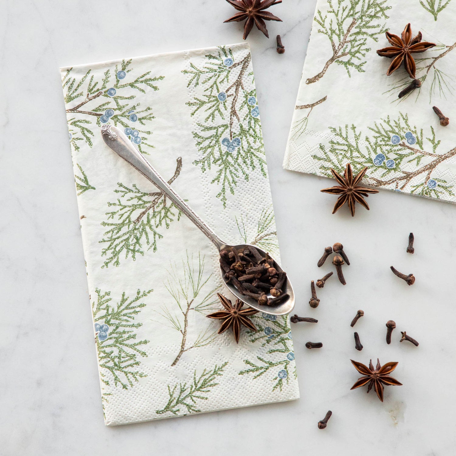 Two Juniper Sprigs Napkins, one Guest and one Cocktail, under a spoon full of cloves with more cloves and star anis scattered over the napkins.