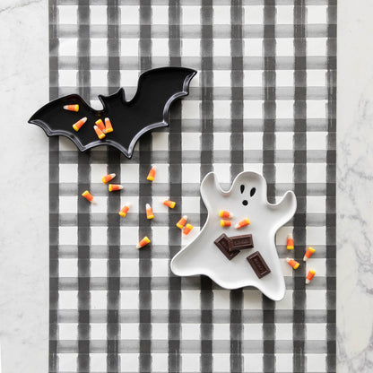 Stoneware Bat Shaped and Ghost Shaped Plates with candy corn and chocolate pieces sprinkled across them, with the Black Painted Check Runner underneath, on a marble table.