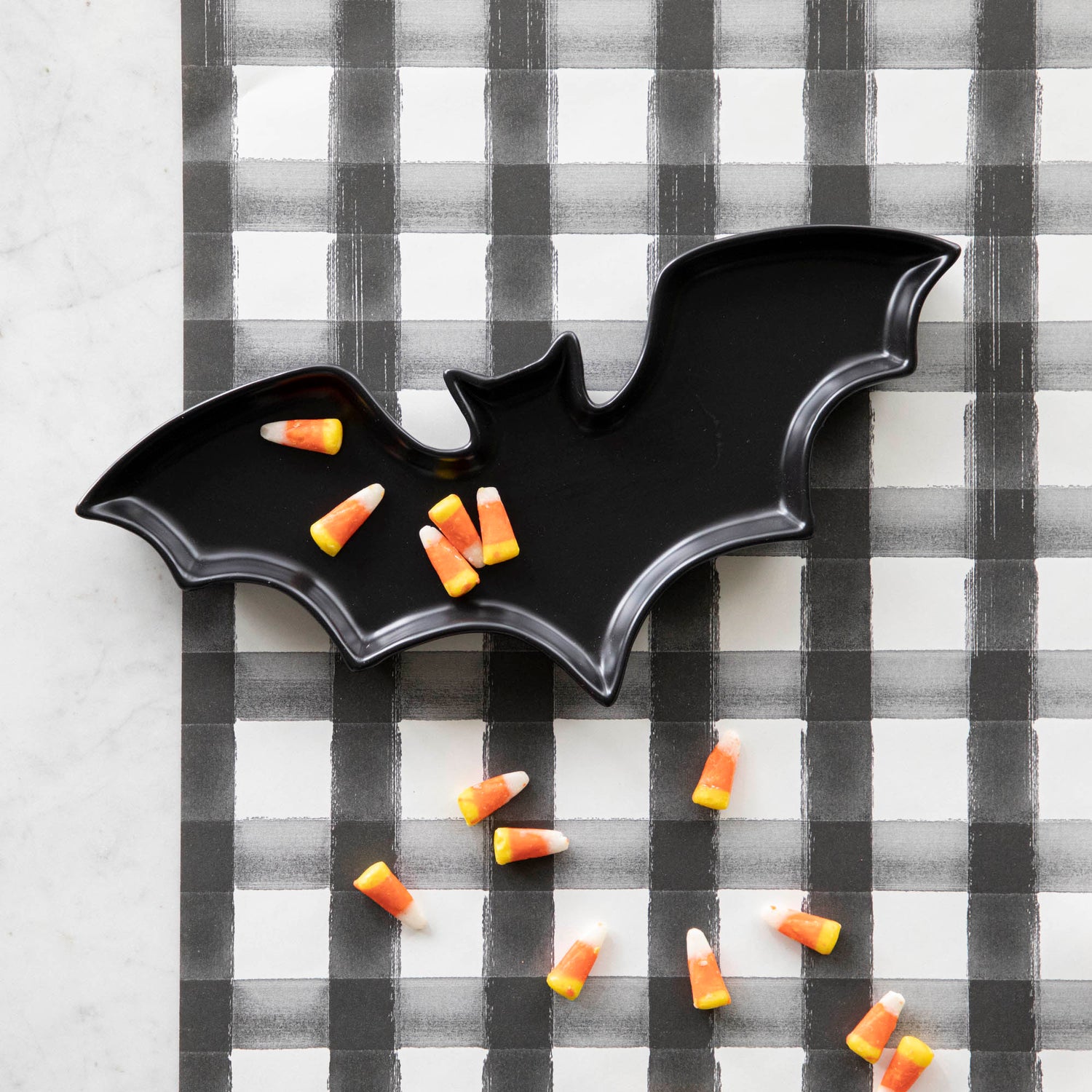 Black Stoneware Bat Shaped Plate with candy corn pieces sprinkled across, and the Black Painted Check Runner underneath.