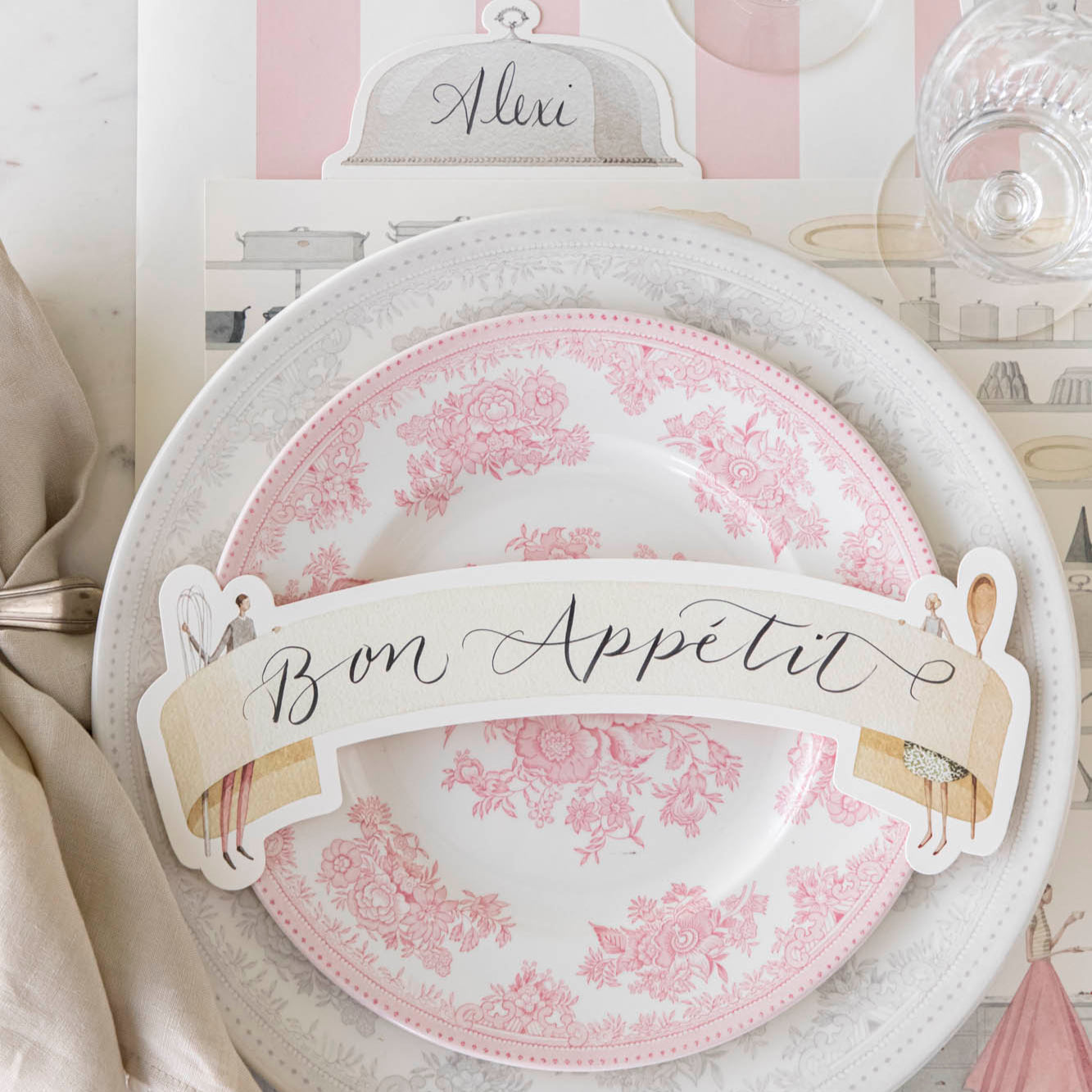 Two Cooks Banner Table Accent with "Bon Appetit" written on it, resting on dinner plates, on an elegant place setting for one.