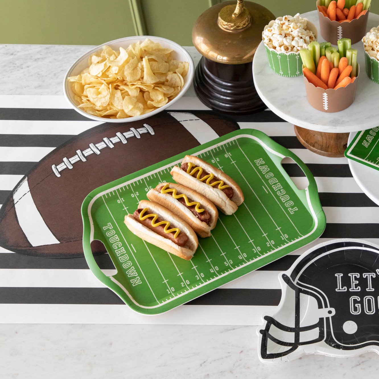 Football field tray of hotdogs on top of a  black stripe runner and die-cut football  placemat, surrounded by various snacks.