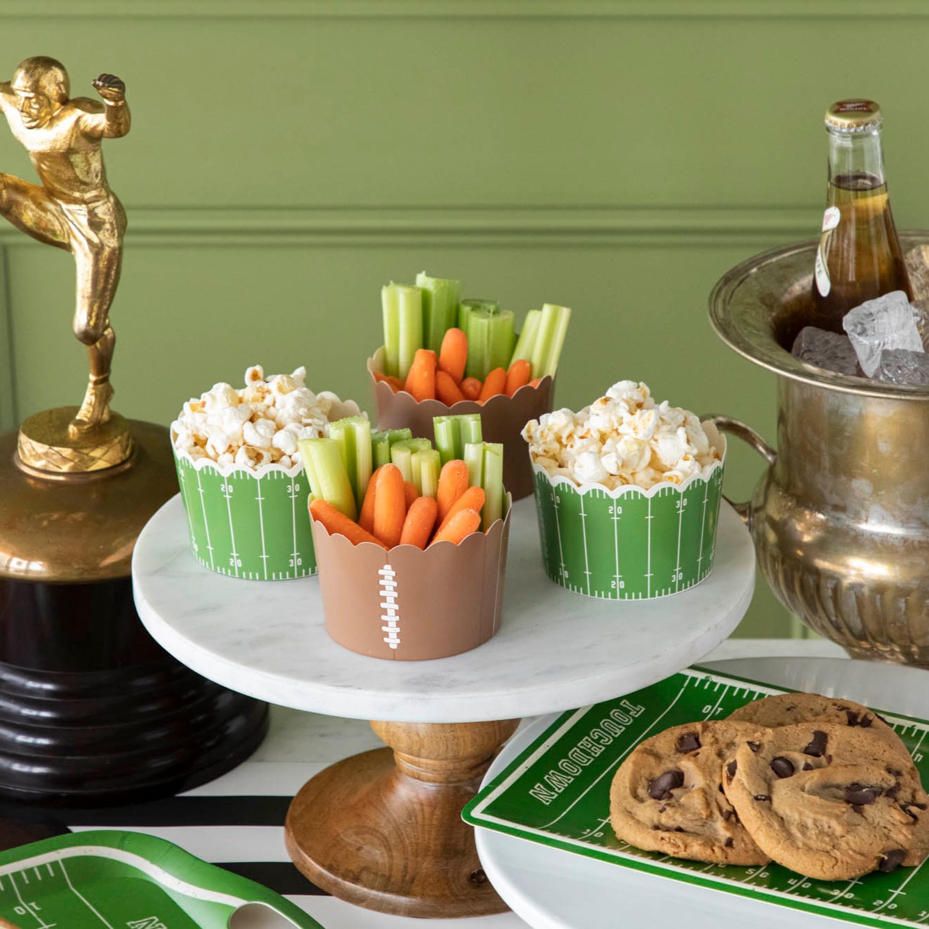 Football field and football paper baking cups filled with vegetables and popcorn on a football themed table set up.