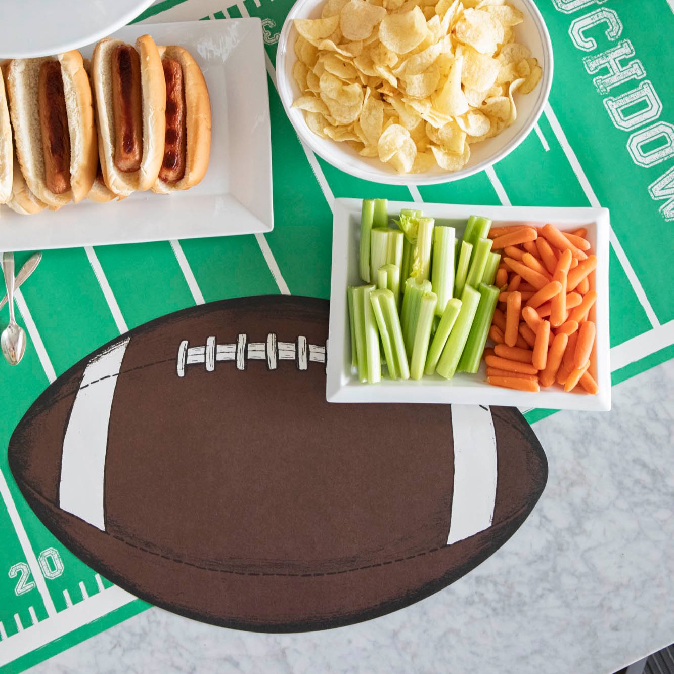 Overhead shot of a die-cut football on top of a touchdown football field runner, with hot dogs, chips and vegetables on various trays.