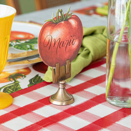 Tomato Place Card with &quot;Mario&quot; written on it, held by the Brass Place Card Holder, behind a place setting.