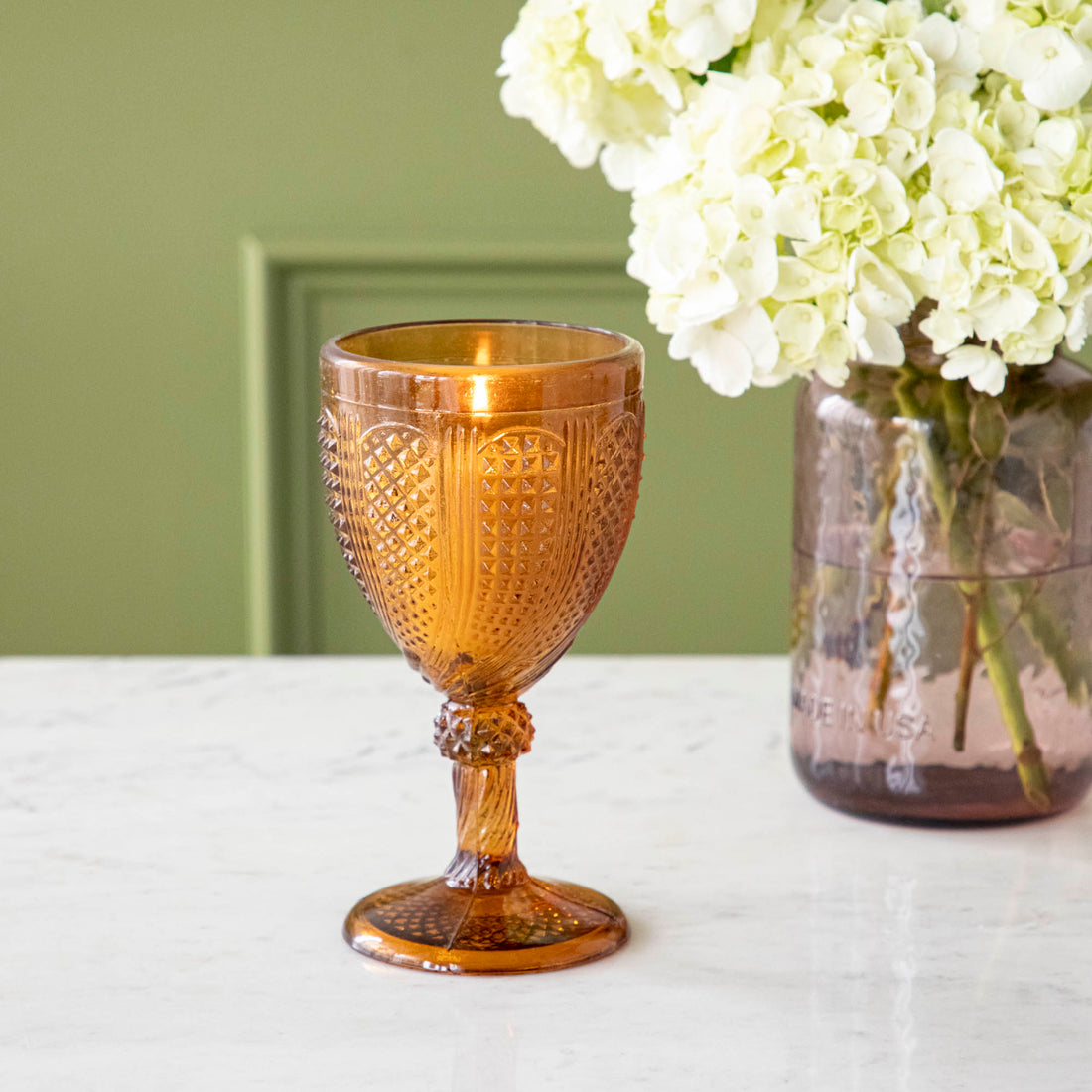 Vintage amber stemmed votive holder with a vase of flowers behind it.