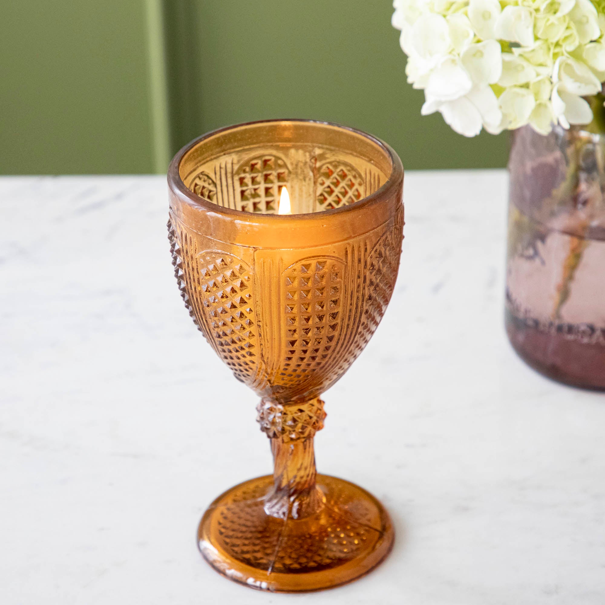 Vintage amber votive holder with a lit candle inside and vase of white flowers behind it.