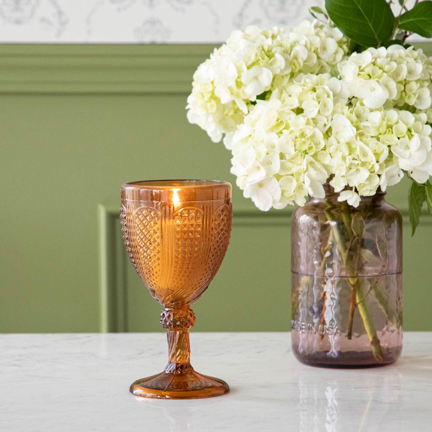 Vintage amber stemmed votive holder with a lit candle inside and a vase of white Hydrangeas in the background.