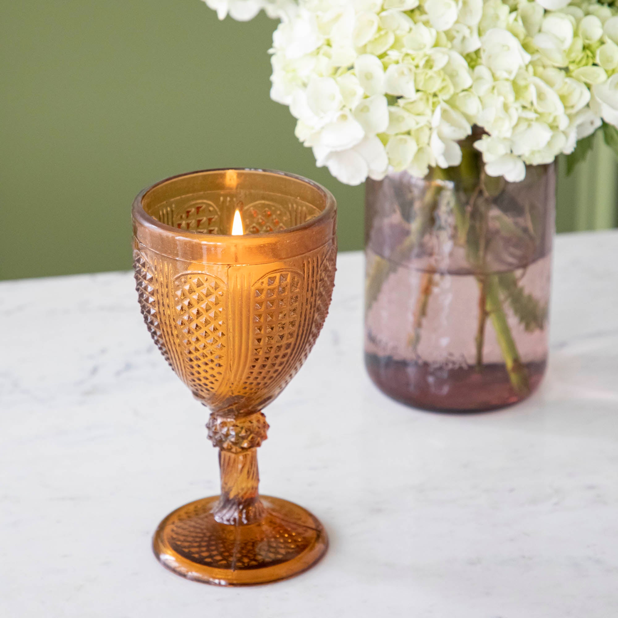 Vintage amber stemmed votive holder with a lit candle inside and a vase of white Hydrangeas in the background.