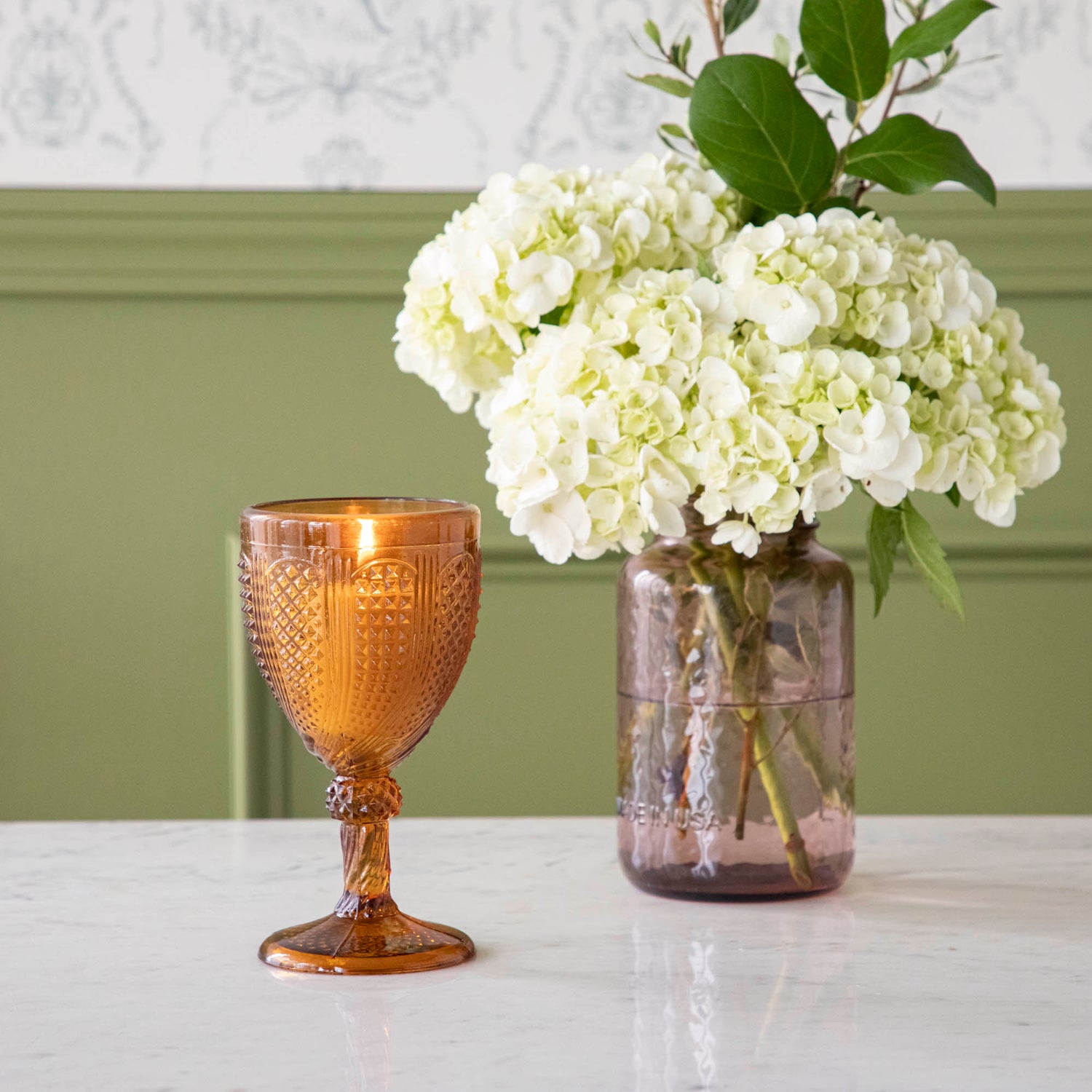 Vintage amber stemmed votive holder with a lit candle inside and a vase of white Hydrangeas in the background.